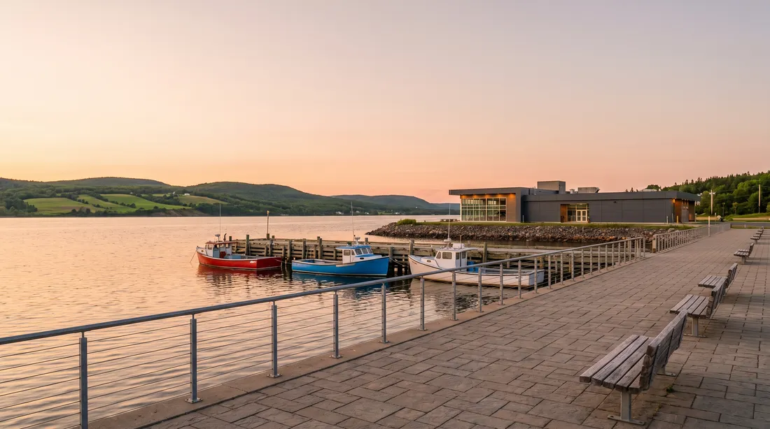 A paved harbourfront walkway with a steel railing and benches at sunset, three small fishing boats at a wooden wharf and green hills across the water.